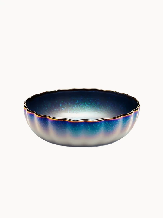 A round ceramic bowl featuring a fluted design with an iridescent, shimmering glaze that gradients from deep metallic blue to white. The artistic dish is highlighted by a delicate gold scalloped rim