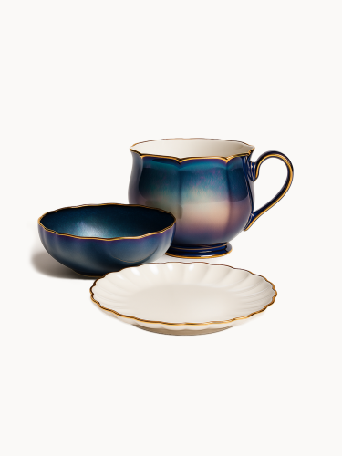 An elegant ceramic breakfast set features a large mug and a matching bowl with an iridescent blue-purple gradient glaze and gold scalloped rims. A white fluted saucer with a matching gold edge sits in the foreground.