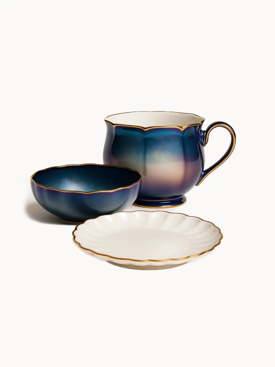 An elegant ceramic breakfast set features a large mug and a matching bowl with an iridescent blue-purple gradient glaze and gold scalloped rims. A white fluted saucer with a matching gold edge sits in the foreground.