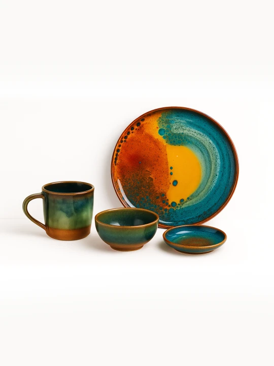 A vibrant ceramic tableware set with a glossy, colorful reactive glaze is arranged against a solid white background. The collection includes a large artistic plate with orange and teal patterns, a matching mug, a medium bowl, and a small saucer.