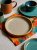 A round ceramic plate with a speckled grey interior and a terracotta rim sits on a vibrant tie-dye cloth. The colorful table setting features an orange mug, a green cup, and turquoise dishes on a speckled countertop.