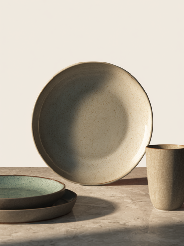 A round ceramic plate featuring a speckled light grey and beige texture with a slightly glossy finish. The minimalist dish is isolated against a plain white background surrounded by matching plates and cups.