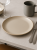A matte, beige ceramic plate sits centered on a light grey countertop. The minimalist table setting includes a grey mug, silver cutlery, and an empty wine glass.