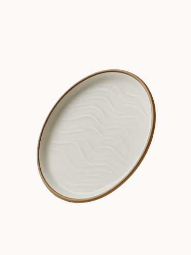 An oval ceramic platter in a matte off-white finish features a textured, wavy sand dune pattern on its interior and a natural brown rim.