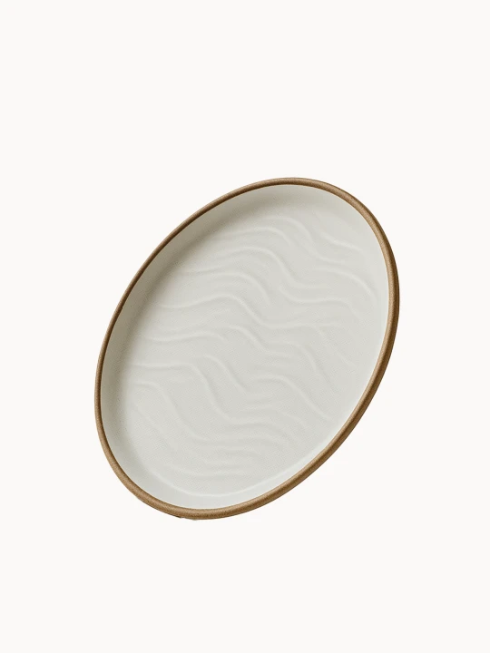 An oval ceramic platter in a matte off-white finish features a textured, wavy sand dune pattern on its interior and a natural brown rim.