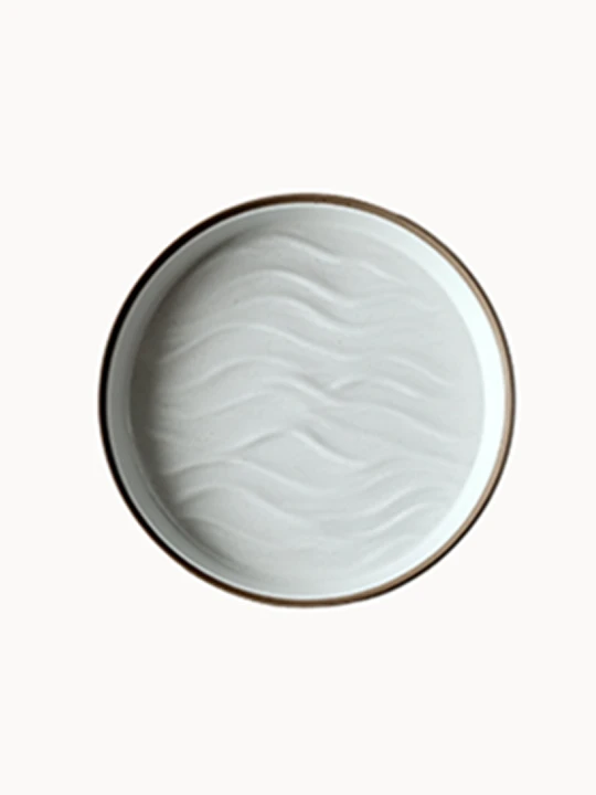 A top-down view of an oval ceramic platter in a matte off-white finish features a textured, wavy sand dune pattern on its interior and a natural brown rim.