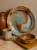 A large ceramic decorative plate with a brown and grey marbled pattern leans against a beige wall on a marble tabletop. The rustic arrangement includes a brown ceramic vase, a speckled mug, a square plate, and a wooden bowl.