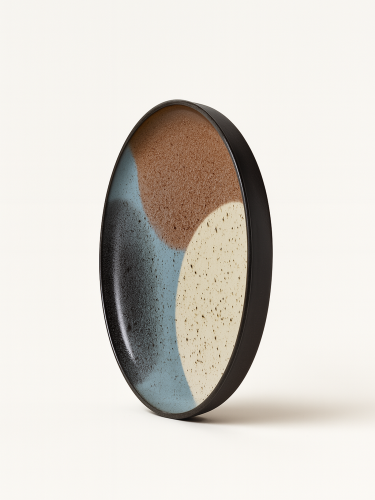 Duży ceramiczny talerz z czarnym rantem oraz  abstrakcyjnym wzorem z w odcieniach brązu, beżu, jasnego błękitu i czerni. Talerz jest zaprezentowany w pozycji pionowej pod lekkim kątem na jednolitym białym tle.