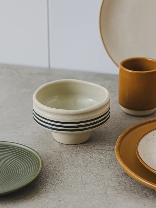 Kremowa ceramiczna miska na podwyższeniu z czarnymi, poziomymi paskami stoi na szarym blacie. Minimalistyczna aranżacja naczyń obejmuje talerze w kolorach ziemi oraz ochrowy kubek w tle.