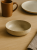 A shallow, speckled beige ceramic bowl is placed on a light wooden table. The minimalist tableware arrangement includes a brown mug, a matching plate, and a linen napkin in the background.