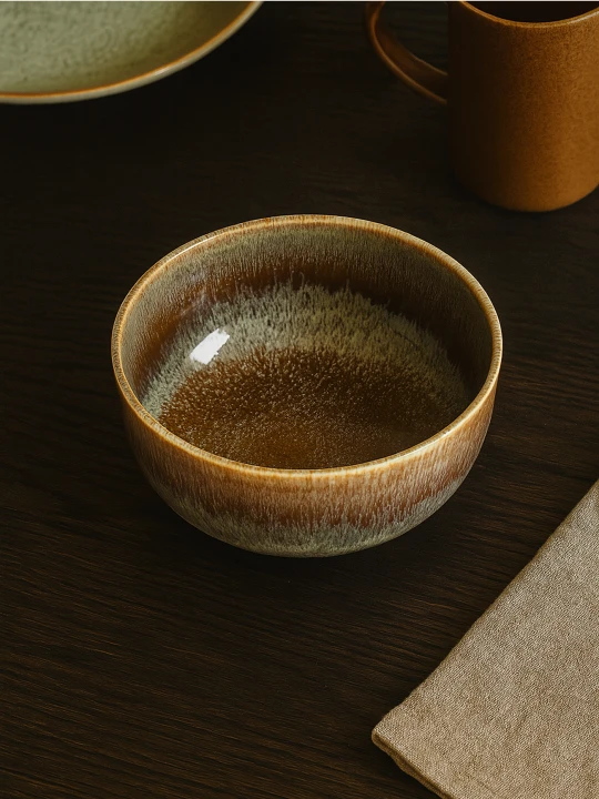 A round ceramic bowl with a dripping brown and beige glaze sits on a dark wooden table. The cozy table setting includes a linen napkin, an ochre mug, and a plate partially visible in the background.