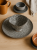 A ceramic bowl with a black and white speckled texture sits on a matching plate on a wooden table. The earthy table setting includes an ochre mug, a grey cup with a saucer, and a linen napkin.