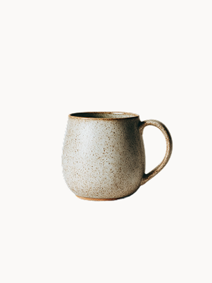A speckled, light beige ceramic mug with a rounded shape and a sturdy handle on a plain white background. The textured surface features fine brown dots, exposing a natural unglazed rim at the very base.