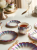 An elegant tea service with iridescent, pearlescent glaze finishes  on a bright white table. Next to a teacup there are matching scalloped plates and gold forks.