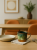 A rustic ceramic mug with a green glaze sits on a light napkin on a wooden table. The cozy table setting includes a ceramic plate and cutlery against a softly blurred dining room background.