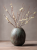 A dark brown, ribbed ceramic vase holding several bare branches with small white plum blossoms sits on a surface. The setting features a rustic, textured table against a solid, warm gray background.