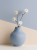 A matte light blue ceramic vase holding a delicate branch with white blossoms is set on a surface. The background features a minimalist composition with intersecting geometric shapes in light blue and beige.