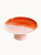 A ceramic cake stand with a two-tone surface, on a white background. The smooth top is half glazed in glossy amber-orange and half in matte pastel pink, supported by a short pink pedestal base.
