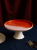 A ceramic cake stand with a two-tone top sits empty on a dark blue tablecloth. The smooth surface is split into glossy orange and matte pale pink, presented next to a gold fork and a partial plate.