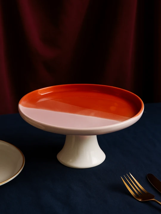 Ceramiczna patera z dwukolorowym blatem na ciemnoniebieskim obrusie. Gładka powierzchnia dzieli się na błyszczący pomarańcz i matowy jasny róż, zaprezentowane obok złotego widelca i fragmentu talerza.