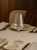 A clear wine glass with a thin stem sits empty on a set table. The setting includes plates and cutlery on a beige tablecloth.