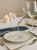 An elegant martini glass in an atmospheric table setting, on a white plate with a gold rim. Silver cutlery and a navy blue napkin sit alongside, with two lit candles in the background.